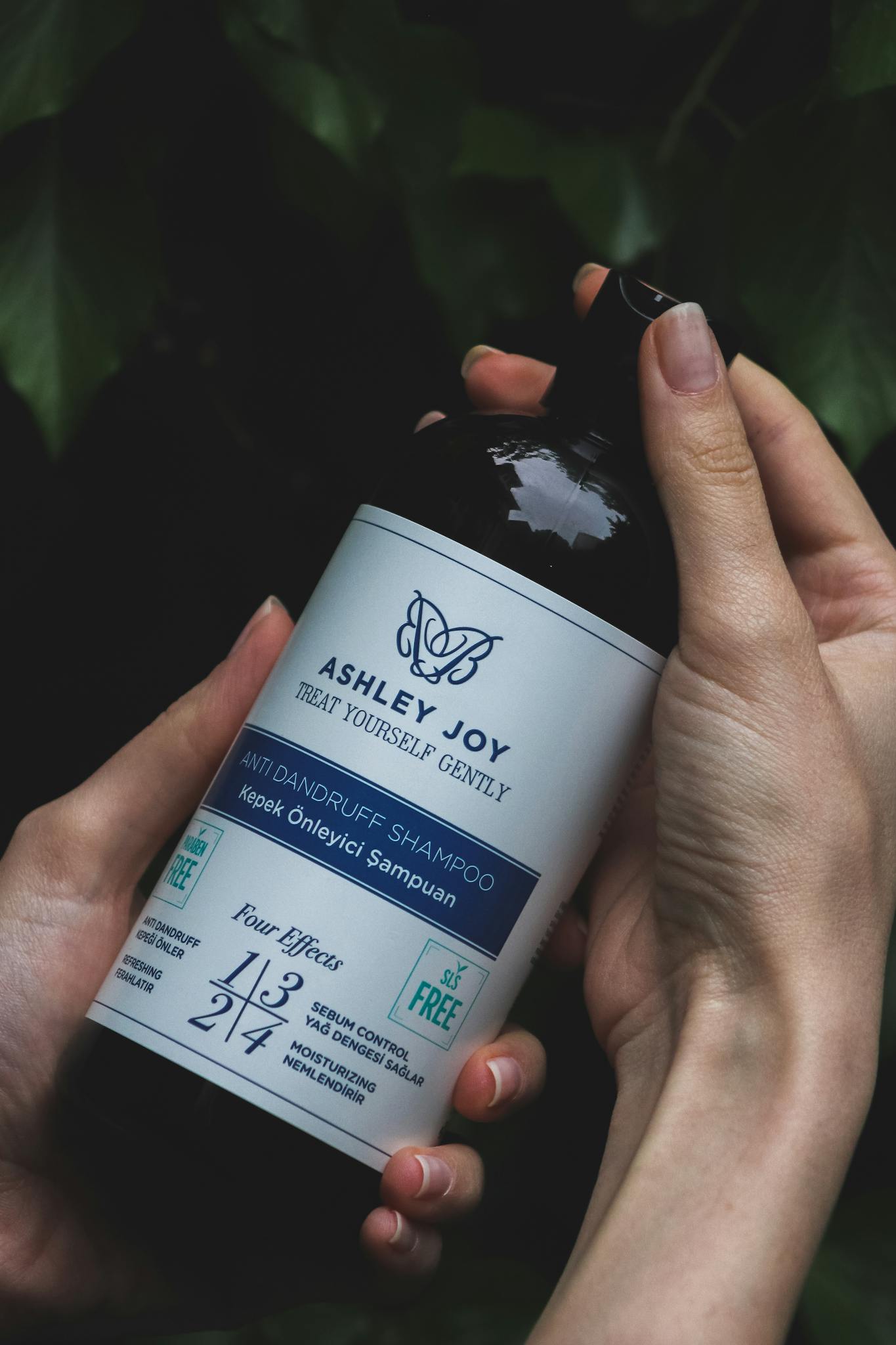 Close-up of hands holding an Ashley Joy anti-dandruff shampoo bottle with nature background.